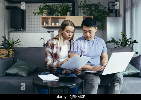 Young multiracial family, Asian man and woman, together at home, focused on home finances, pay bills online, using a laptop Stock Photo