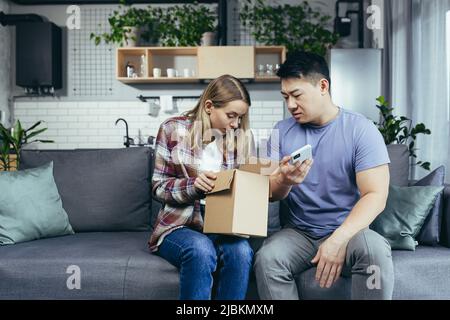 Multiracial family at home on the couch disappointed to receive a parcel, cheated in an online store, a man and a woman sitting on the couch Stock Photo