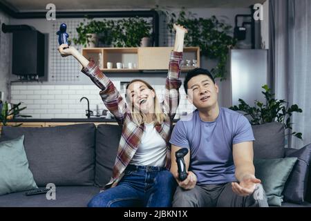 Happy couple spending leisure time together at home, playing computer games, holding joysticks, happy multiracial family Stock Photo