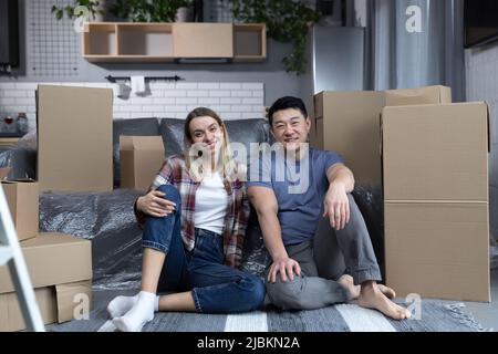 Young couple man and woman hugging together happy looking at camera, sitting on the floor of a new apartment, happy multiracial family Stock Photo