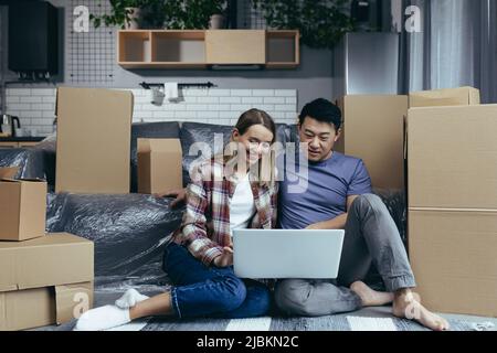 Loving and happy couple sitting on the floor, Asian man and woman among cardboard boxes, looking at laptop screen, shopping online in online store Stock Photo