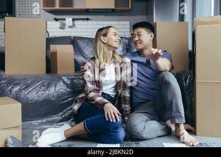Close-up photo of a young family in a new apartment holding the keys to the house, a multiracial couple sitting in the living room among the cardboard Stock Photo