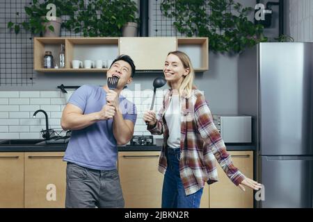Happy young Asian man and woman couple singing and dancing together in the kitchen, having fun together, family in love Stock Photo