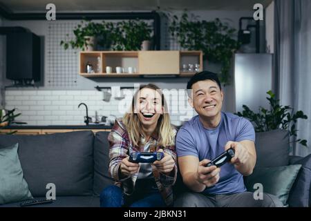 Happy couple spending leisure time together at home, playing computer games, holding joysticks, happy multiracial family Stock Photo