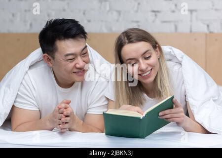 Happy Asian couple in love and woman lying together in bed and reading a book Stock Photo