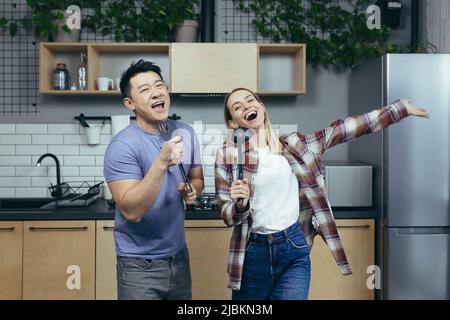 Happy young Asian man and woman couple singing and dancing together in the kitchen, having fun together, family in love Stock Photo