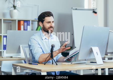 Successful and happy business coach mentor, records audio podcast, uses professional microphone, businessman in modern white office, conducts online b Stock Photo