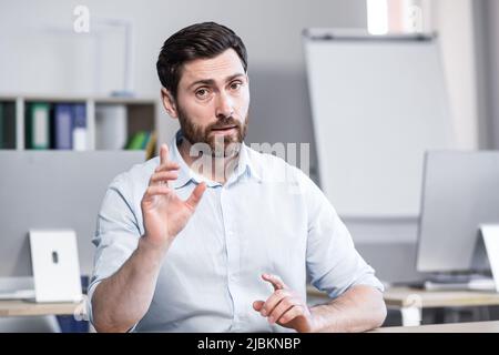 Young handsome successful business coach explains and tells on camera, gestures with his hands. Sitting at a computer desk in the office, looking at t Stock Photo