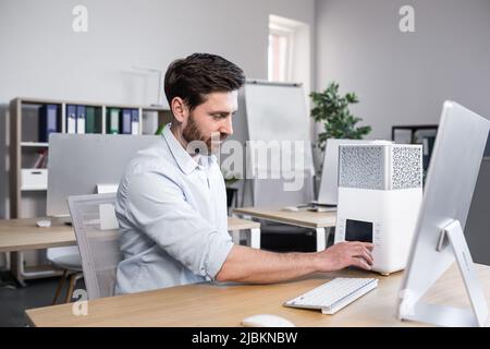 Young handsome man, office worker, works includes humidifier, air conditioner which stands on the table near the computer in the office Stock Photo
