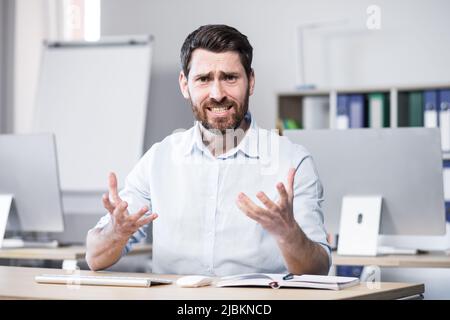 Young handsome successful business coach explains and tells on camera, gestures with his hands. Sitting at a computer desk in the office, looking at t Stock Photo