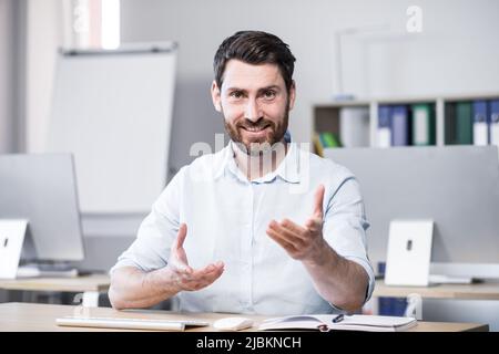 Young handsome successful business coach explains and tells on camera, gestures with his hands. Sitting at a computer desk in the office, looking at t Stock Photo