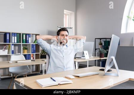 Young handsome man, office worker, sitting at a computer desk, stretching, tired, taking a break, doing yoga exercises, resting Stock Photo