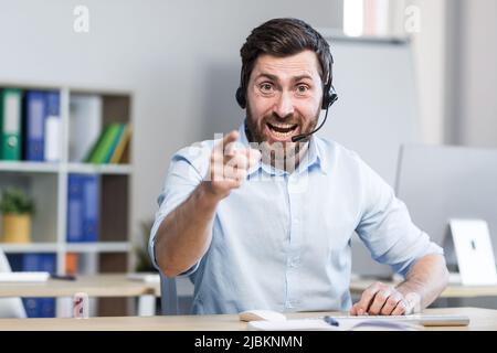 A young man, a consultant, an office worker, a manager talking on a video call in headphones, waving his arms, explaining something. Sitting at a comp Stock Photo