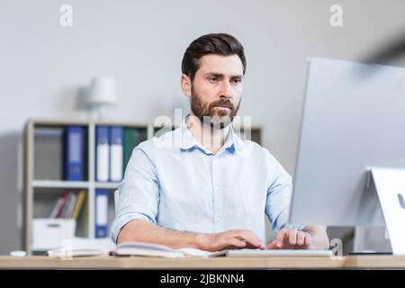 Portrait of a young man, focused and serious office worker, manager, director working at a computer, sitting behind a desk in the office Stock Photo