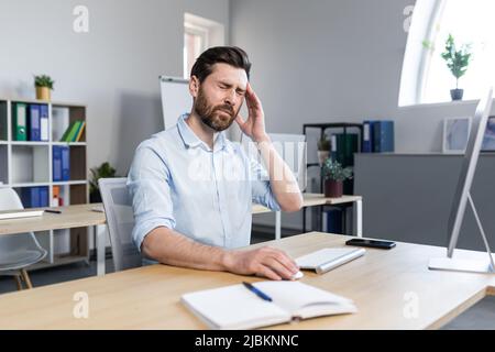 The man works in the office, the patient has a severe headache, upset businessman sitting at the table, holding his hands behind his head Stock Photo
