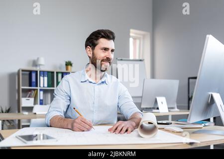 Young male architect, designer making drawings and project on whatman paper on desk in his office. Sitting at a table, with a computer, focused Stock Photo