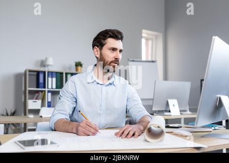 Young male architect, designer making drawings and project on whatman paper on desk in his office. Sitting at a table, with a computer, focused Stock Photo