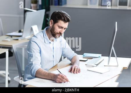 Young male architect, designer making drawings and project on whatman paper on desk in his office. Sitting at a table, with a computer, focused Stock Photo