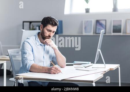 Young male architect, designer making drawings and project on whatman paper on desk in his office. Sitting at a table, with a computer, focused Stock Photo