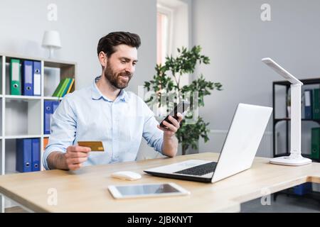 Successful online shopping through a smartphone. Successful deal. Satisfied young man holds a credit card and a mobile phone, makes an order. Sitting Stock Photo