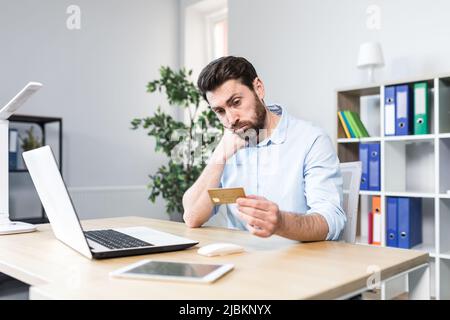 Unsuccessful online purchase. Unsuccessful deal. Upset young man, office worker holding a credit card, holding his head in shock. Sitting at a desk, a Stock Photo