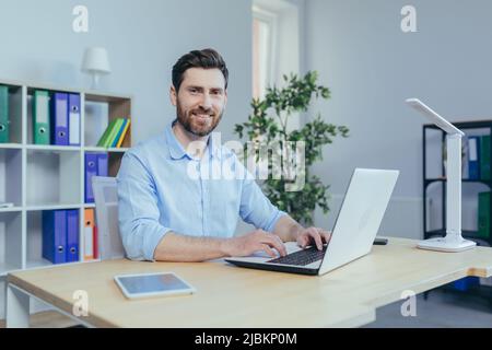 Happy and successful man working on laptop in home office, sitting at table in bright room, smiling looking at camera Stock Photo
