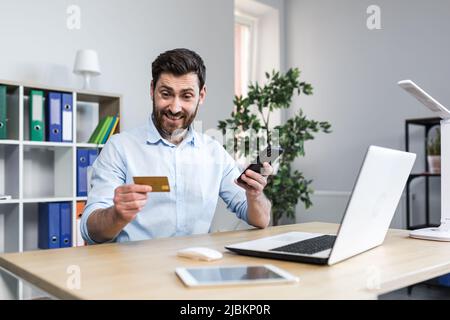 Successful online shopping through a smartphone. Successful deal. Satisfied young man holds a credit card and a mobile phone, makes an order. Sitting Stock Photo