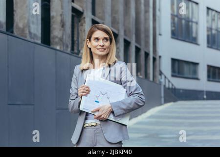 Happy and successful business woman with laptop and documents outside office smiling in business suit Stock Photo