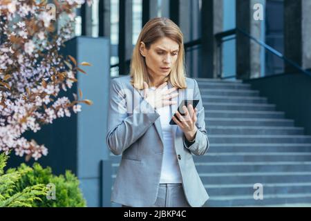 Frustrated and shocked business woman reading bad news online from phone, businesswoman outside office in business suit Stock Photo