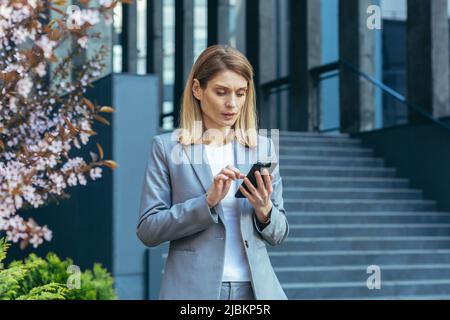 Serious and pensive business woman uses the phone outside the office, looking at the smartphone screen Stock Photo
