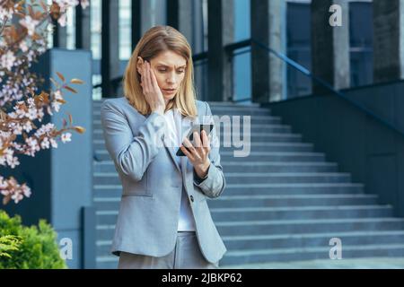 Frustrated and shocked business woman reading bad news online from phone, businesswoman outside office in business suit Stock Photo