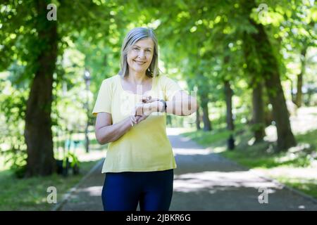 Active lifestyle. Senior beautiful woman running in the park in the morning. Looks and monitors the pulse on the watch on his hand. Goes in for sports Stock Photo