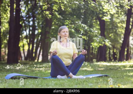 Senior gray-haired retired woman in the park meditates and performs breathing exercises, summer day in lotus position Stock Photo