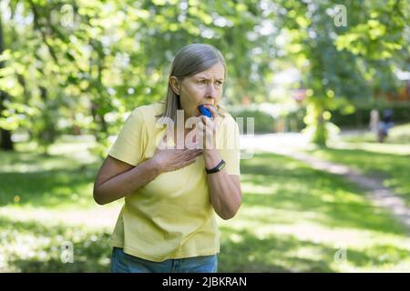 Senior retired woman with asthma breathing in an inhaler in a summer park Stock Photo