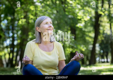 Senior gray-haired retired woman in the park meditates and performs breathing exercises, summer day in lotus position Stock Photo