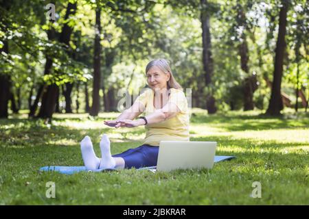 Active gray-haired woman in the park doing fitness sitting on the mat and grass, using a laptop for online classes in the park Stock Photo