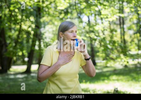 Senior retired woman with asthma breathing in an inhaler in a summer park Stock Photo