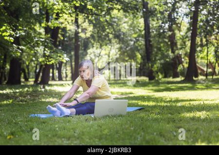 Active gray-haired woman in the park doing fitness sitting on the mat and grass, using a laptop for online classes in the park Stock Photo