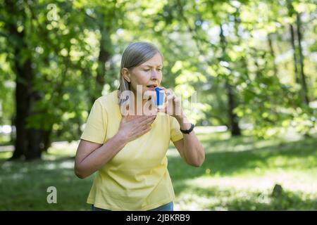 Senior retired woman with asthma breathing in an inhaler in a summer park Stock Photo