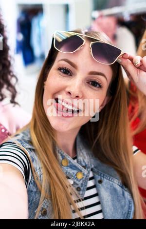 Smiling girlfriends wearing stylish sunglasses having fun time taking selfie with mobile phone while doing shopping in clothing store. Stock Photo