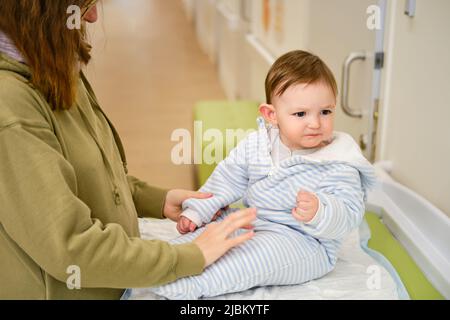 Mom with a child in the corridor of the hospital on the table for changing clothes. Mother with toddler boy on changing table in clinic lobby. One yea Stock Photo