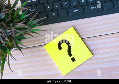 Question mark on adhesive note on wooden desk with black keyboard and potted plant. Stock Photo