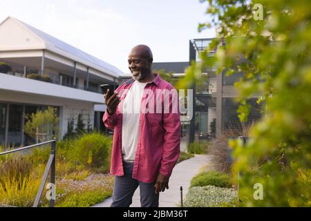 Smiling bald african american senior man using smartphone while standing against house in yard Stock Photo