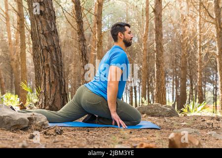 Handsome young man practicing yoga stretching at sunset in the forest surrounded by trees on his blue mat. Stock Photo