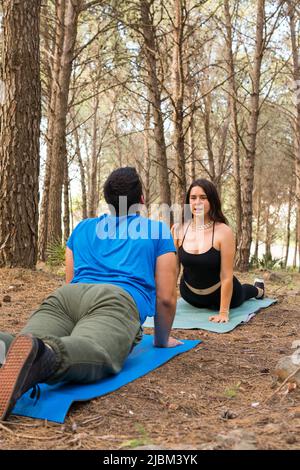 Friends practicing yoga facing each other at sunset in a forest with trees around them with their mats on the ground. Stock Photo