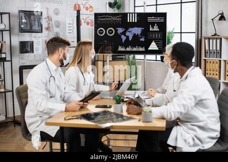 Multiethnic employees in protective masks working with tablet and laptops during meeting in hospital boardroom. Doctors analyzing spread of coronavirus while looking at monitor with researches. Stock Photo