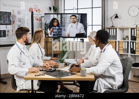 Group of four multicultural physicians watching online lecture about MRI diagnosis in modern hospital. Confident doctors analyzing ways of successful treatment during video conference with colleagues. Stock Photo