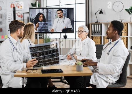 Teamwork of medical scientists examining x ray of patient lungs in modern clinic. Multicultural group of physicians in lab coats discussing MRI result with two colleagues during online video call. Stock Photo