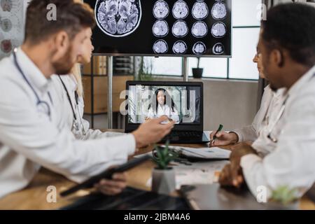Diverse team of medical scientist having online meeting with african american speaker about brain anomalies in high tech laboratory. Colleagues using laptop while sitting at table in neurology center. Stock Photo
