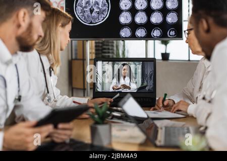 Diverse team of medical scientist having online meeting with african american speaker about brain anomalies in high tech laboratory. Colleagues using laptop while sitting at table in neurology center. Stock Photo